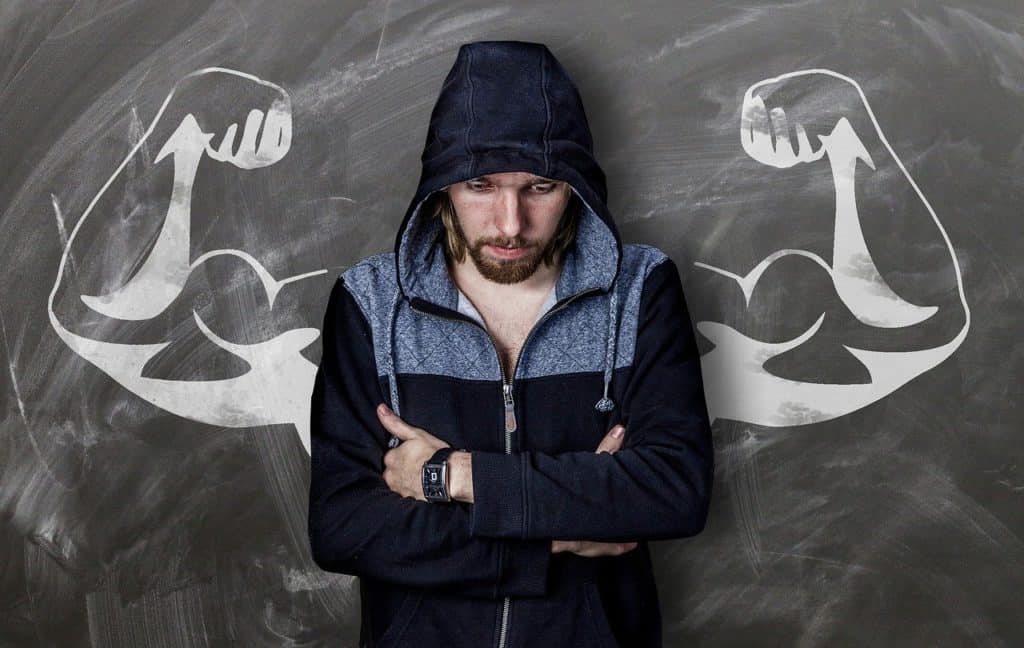 A man with his arms crossed in front of him standing against a wall between two muscular arms drawn on a wall to signify the difference in strength building between Pilates vs. yoga